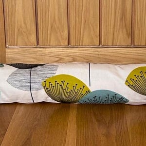 May include: A long, rectangular door draft stopper with a matching smaller piece. The fabric is off-white with a repeating pattern of stylized yellow, teal, and gray floral shapes. The stopper is resting on a wooden floor in front of a wooden door.
