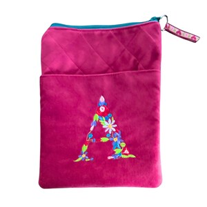 May include: A magenta plush crossbody bag featuring an embroidered floral "A". The bag has a quilted top section, a teal zip, and a small strap with heart detailing. The bag is designed to be worn over the shoulder, with a length of approximately 25 cm.