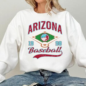 May include: White sweatshirt with a baseball-themed graphic. The design features the word "ARIZONA" in red, with "19" and "98" on either side of a baseball diamond graphic. The word "Baseball" is written in a curved font.