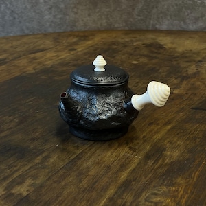 May include: A small, black ceramic teapot with a textured surface. The teapot has a white handle and a white finial on the lid. The spout has a small red accent. The teapot is sitting on a wooden surface.