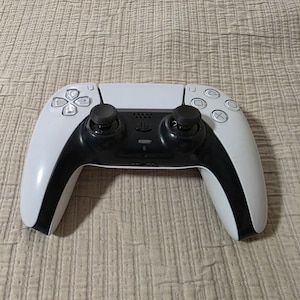 May include: A white and black wireless video game controller featuring a central touchpad, two black analog sticks, a directional pad, and four action buttons marked with triangle, circle, square, and cross symbols on the right side.