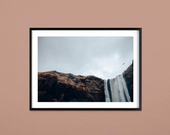 Pode incluir: Fotografia de paisagem emoldurada de uma cachoeira em cascata por um penhasco rochoso. A imagem apresenta um céu nublado e um pássaro em voo. A cachoeira é o ponto focal, com água fluindo pelo centro da moldura.