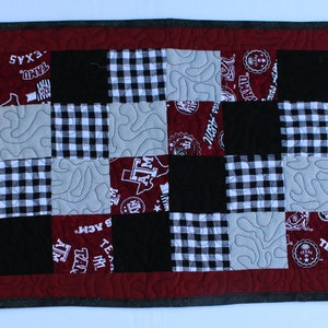 May include: A rectangular quilted table runner with a patchwork design. The runner features alternating squares of black, white gingham, gray, and maroon fabric. The maroon squares have white text and logos. The border is maroon with a gray edge.