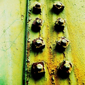 May include: Close-up of a rusty metal surface with a row of square-shaped metal rivets. The rivets are evenly spaced and have a worn, aged appearance. The surface is a mix of green and yellow, with patches of rust.
