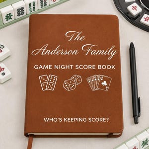 Puede incluir: Un cuaderno marrón con texto blanco que dice The Anderson Family GAME NIGHT SCORE BOOK y WHO'S KEEPING SCORE? La portada presenta dibujos lineales blancos de dos fichas de dominó, dos dados y cinco cartas de juego con un bolígrafo negro.