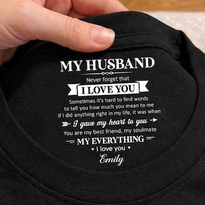 May include: A black t-shirt featuring white printed text on the inside collar area that reads: MY HUSBAND, Never forget that I LOVE YOU, Sometimes it's hard to find words to tell you how much you mean to me, I gave my heart to you, MY EVERYTHING, Emily.