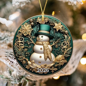 May include: A round Christmas ornament featuring a central snowman wearing a green top hat and a gold scarf. The design includes intricate gold filigree, holly leaves, and stars against a dark green background with the year 2026 printed at the bottom.