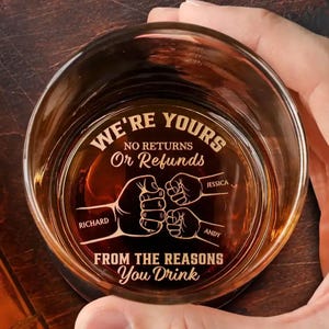 May include: A clear whiskey glass with the text "WE'RE YOURS" and "FROM THE REASONS You Drink" etched on the bottom. The glass features a design of four fists bumping, with the names Richard, Jessica, and Andy. The glass is filled with a brown liquid.