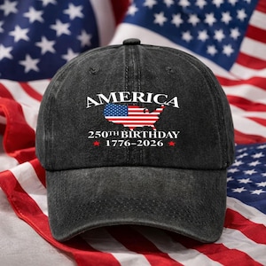 Puede incluir: Una gorra de béisbol de mezclilla negra con la palabra "AMERICA" sobre un mapa de los Estados Unidos, coloreado con la bandera estadounidense. El texto "250TH BIRTHDAY 1776-2026" está debajo. Banderas americanas en el fondo.