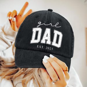 Puede incluir: Gorra de béisbol negra con las palabras "girl DAD EST. 2025" en blanco. La gorra está sujeta por una mano. La gorra es de estilo clásico.