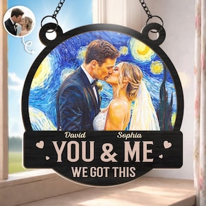 Puede incluir: Una placa de madera circular colgante que muestra a una pareja con traje y vestido de novia sobre un fondo azul estrellado. Los nombres David y Sophia están impresos sobre el texto YOU & ME WE GOT THIS, con pequeños corazones.