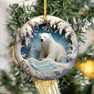 Puede incluir: Un adorno navideño redondo de cerámica con una ilustración en estilo 3D de un oso polar blanco sentado en un paisaje invernal nevado con pinos y un cielo azul. El año 2026 está impreso en color negro en la parte inferior central.