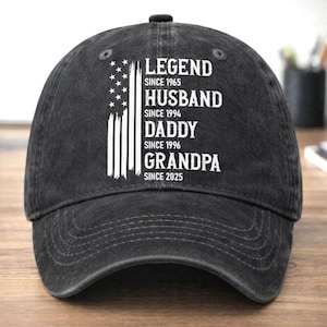 May include: A black baseball cap with white text and a distressed American flag design. The text reads "LEGEND SINCE 1965, HUSBAND SINCE 1994, DADDY SINCE 1996, GRANDPA SINCE 2025". The cap has a curved brim and a black button on top.