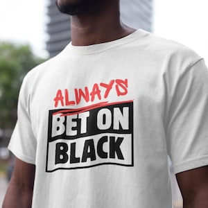 May include: White t-shirt with the text "ALWAYS BET ON BLACK" in red and black. The text is in a bold, blocky font, with "ALWAYS" and "BET ON" in a rectangular box. The shirt is a classic crew neck style.