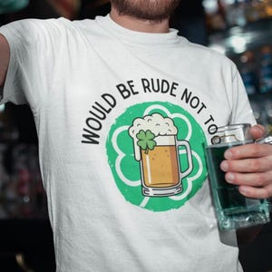 Może przedstawiać: Biała koszulka z zieloną koniczyną i grafiką kufla piwa. Tekst na koszulce brzmi "WOULD BE RUDE NOT TO". Osoba trzyma szklany kufel wypełniony zielonym płynem.