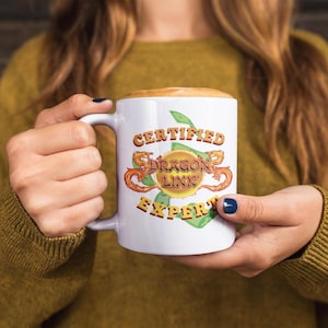 Könnte beinhalten: Weiße Keramik-Tasse mit dem Schriftzug "CERTIFIED DRAGON LINK EXPERT" in Orange und Gelb. Das Design umfasst eine Goldmünze mit Drachen und grünen Blättern. Die Tasse wird von einer Person gehalten.