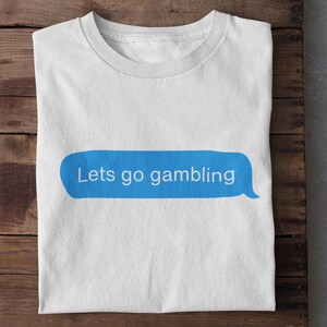 May include: White t-shirt with a blue text bubble graphic that reads "Lets go gambling" in a sans-serif font. The shirt is folded and placed on a wooden surface.