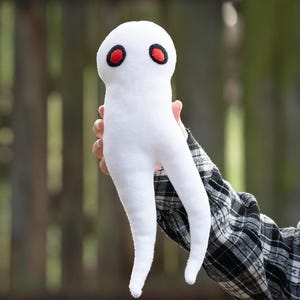 Może przedstawiać: Biała pluszowa zabawka w kształcie postaci przypominającej ducha z czerwonymi oczami. Miękka zabawka jest trzymana na tle rozmytego tła zewnętrznego. Postać ma zaokrągloną głowę i dwie długie, zwisające nogi.