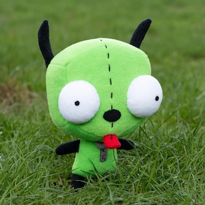K&ouml;nnte beinhalten: Ein gr&uuml;nes Pl&uuml;sch-Alien-Puppe mit gro&szlig;en wei&szlig;en Augen, schwarzen Antennen und einer roten Zunge. Die Puppe hat schwarze Arme und F&uuml;&szlig;e und ein kleines graues Rei&szlig;verschlussdetail auf der Brust. Sie steht auf gr&uuml;nem Gras.