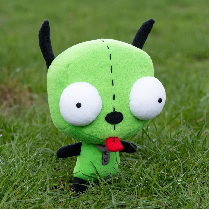 K&ouml;nnte beinhalten: Ein gr&uuml;nes Pl&uuml;sch-Alien-Puppe mit gro&szlig;en wei&szlig;en Augen, schwarzen Antennen und einer roten Zunge. Die Puppe hat schwarze Arme und F&uuml;&szlig;e und ein kleines graues Rei&szlig;verschlussdetail auf der Brust. Sie steht auf gr&uuml;nem Gras.