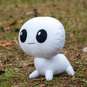 May include: A white plush toy with a round head and large black eyes. The toy has a simple, cartoon-like design and is sitting on a bed of brown grass.