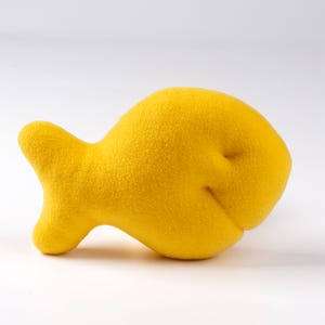 Puede incluir: Un juguete de peluche de pescado amarillo. El pez está hecho de tela suave y peluda y tiene una forma ligeramente curvada.