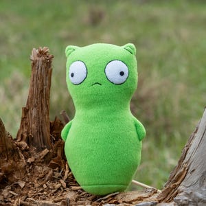 Op de afbeelding: Een groene pluche speelgoed dat lijkt op een alien met grote witte ogen en kleine zwarte pupillen. Het zachte speelgoed heeft een eenvoudig ontwerp met kleine armen en oren. De achtergrond is een vage buitenscène met een boomstronk.
