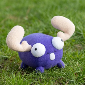 May include: A plush purple toy with large white eyes and a toothy grin. The toy has two large, cream-colored antlers.