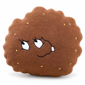 Puede incluir: Un cojín de peluche marrón con forma de galleta y superficie suave y texturizada. La galleta presenta detalles bordados que incluyen ojos blancos con pupilas negras, pestañas negras, una pequeña boca negra y puntadas amarillas dispersas.