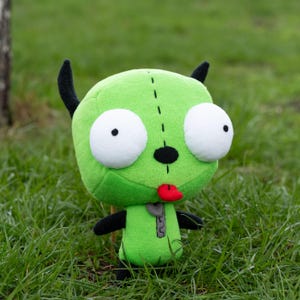 K&ouml;nnte beinhalten: Ein gr&uuml;nes Alien-Pl&uuml;schtier mit gro&szlig;en wei&szlig;en Augen, schwarzen H&ouml;rnern und einer roten Zunge. Die Puppe hat eine schwarze Nase und einen Rei&szlig;verschluss auf der Brust. Es steht im gr&uuml;nen Gras.