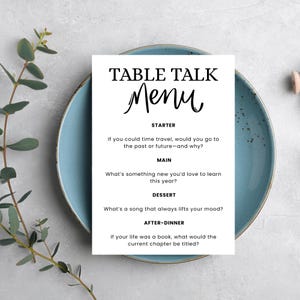 May include: A white "Table Talk Menu" card with black text, including "Menu" in cursive, sits on a light blue plate. The menu features conversation starters for a meal, such as "If you could time travel..." and "What's a song that lifts your mood?"