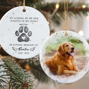May include: A round, white ceramic ornament with a gold string. One side features a watercolor image of a golden retriever. The other side has text: "NO LONGER BY MY SIDE FOREVER IN MY HEART" and "IN LOVING MEMORY OF Barten 2021-2025" with a paw print.