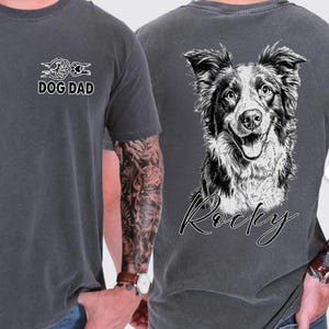 May include: A charcoal grey t-shirt featuring a small graphic on the front chest with a fist bumping a paw and the text DOG DAD. The back of the shirt displays a large, detailed black and white sketch of a dog's face with the name Rocky below it.