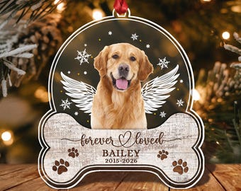 May include: A memorial ornament shaped like a dog bone with a golden retriever portrait featuring white angel wings and snowflakes. The text reads forever loved, BAILEY, and 2015-2026, accented with paw prints on a light wood grain background.