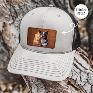 May include: A light gray and black trucker hat featuring a rectangular brown patch on the front. The patch displays a printed image of an orange cat and a black and white dog sitting together. A white circular graphic with the text Printed Patch points to it.