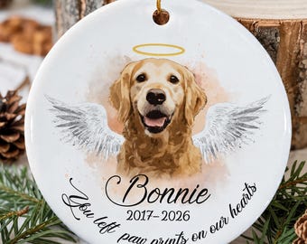 May include: A round white ceramic ornament featuring a watercolor illustration of a golden retriever with white angel wings and a gold halo. The text reads Bonnie, 2017-2026, and You left paw prints on our hearts in black script.