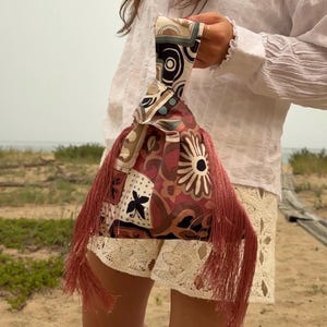May include: A fabric wristlet bag featuring a multi-colored geometric and floral pattern in shades of pink, brown, cream, and black. The bag has a knotted handle and long, dangling pink fringe tassels on both sides of the main pouch body.