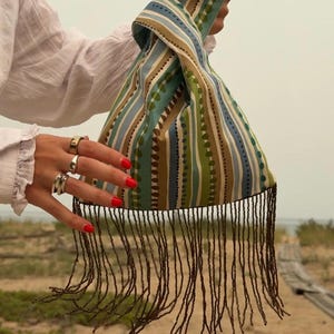 May include: A fabric knot bag featuring vertical stripes in shades of olive green, light blue, and tan, accented by a dotted line pattern. The bottom edge of the bag is finished with long, thin, dark brown beaded fringe that hangs downward.