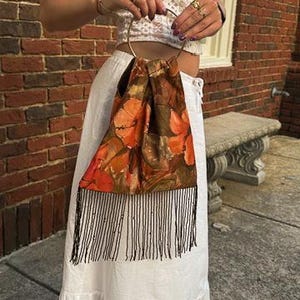 May include: A fabric handbag featuring a vibrant orange and brown floral pattern with a circular gold-tone metal handle and a long fringe trim along the bottom edge. The bag has a gathered top construction and hangs from a single rigid metal ring.