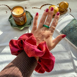 May include: A large, red scrunchie with a textured appearance is held in a hand with red nail polish and a green ring. A lit candle in a brown jar sits on a green dish. A copy of Paris Review is visible.
