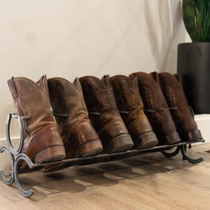 Puede incluir: Un zapatero de metal con seis pares de botas de vaquero marrones. El zapatero está hecho de herraduras y está diseñado para sostener las botas en posición vertical.