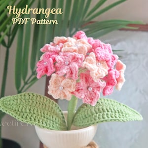 Könnte beinhalten: Ein handgefertigtes Häkel-Hortensien-Blumenarrangement in einer weißen Vase. Die Blume hat rosa und pfirsichfarbene Blütenblätter mit Perlenakzenten, grüne Blätter und einen grünen Stiel. Die Vase ist mit Bindfaden gebunden. Der Text auf dem Bild lautet "Hydrangea PDF Pattern".