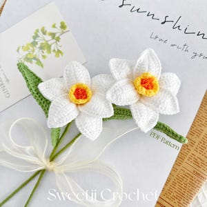 Könnte beinhalten: Zwei gehäkelte weiße Narzissen mit gelben und orangen Zentren, die mit einem weißen Band und grünen Stielen zusammengebunden sind. Die Blumen befinden sich auf einem weißen Hintergrund mit dem Text "Love with you" und "Sunshine" in Schwarz.
