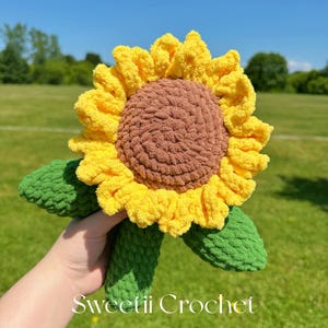 Puede incluir: Un girasol de ganchillo hecho a mano con un centro marrón, pétalos amarillos y hojas verdes. El texto "Sweetii Crochet" es visible en la parte inferior. El girasol se sostiene sobre un fondo de hierba verde y cielo azul.