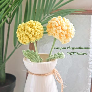 May include: A white ceramic vase tied with twine holds two handmade crochet flowers. One is yellow, the other is a lighter shade of yellow. The flowers have green stems and leaves. The text "Pompon Chrysanthemum PDF Pattern" is visible.