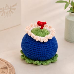 May include: A round, crocheted plush toy featuring a deep blue base, a light green top, and a ruffled cream-coloured border. A small, bright red sprout sits at the centre of the green top, and a scalloped light green base completes the design.