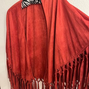 May include: A rust-colored suede fringed shawl. The open-front shawl is hanging on a zebra-print hanger. The shawl has long fringe along the bottom edge.