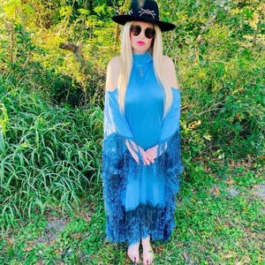 May include: A woman wearing a black hat with a silver design, sunglasses, and a blue dress with fringe. The dress has a high neck and cold shoulder design. The dress transitions from light blue to dark blue with fringe at the bottom.