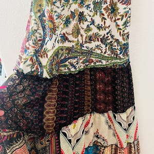 May include: A long, flowing dress with a patchwork design. The dress features a variety of patterns, including floral, paisley, and geometric prints in a mix of colors such as white, blue, red, and brown. The dress has a tiered construction with ruffled edges.