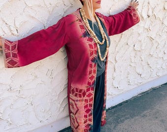 May include: A long, flowing, burgundy kimono-style robe with geometric patterns and fringe detailing. The robe is paired with a gray top, dark blue pants, and light-colored sandals. A burgundy head wrap, sunglasses, and a wooden bead necklace complete the bohemian look.